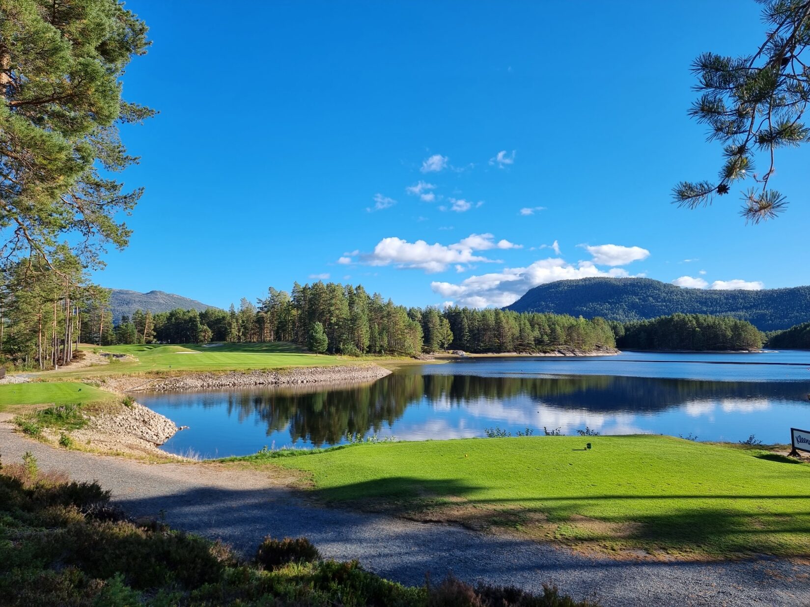 Hull 1 på Vrådal Golf med grønt utslag, blankt vann og fjell under blå sommerhimmel